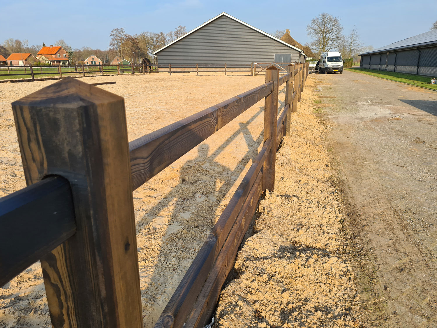 Een manege wordt omzoomd door een elegant paardenhek. Op de achtergrond staan verschillende stalgebouwen.